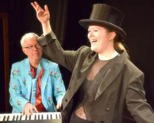 Eine fröhliche Aufführung mit einer Sängerin in einem eleganten Outfit und Zylinder. Im Hintergrund spielt ein Musiker am Klavier.