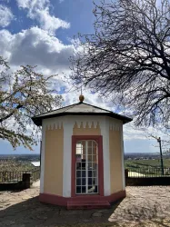 Ein kleiner Pavillon auf einem Hügel, umgeben von Bäumen und mit Blick ins Tal. Der Himmel ist teilweise bewölkt und die Landschaft ist grün.