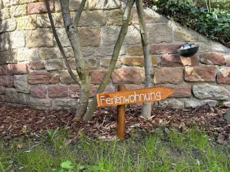 Ein Holzschild mit der Aufschrift "Ferienwohnung" zeigt in Richtung des Weges. Im Hintergrund ist eine Steinmauer und ein kleiner Garten sichtbar.