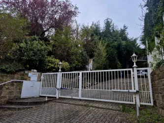 Ein Eingang mit einem weißen Tor, das von grünen Pflanzen umgeben ist. Im Hintergrund sind Bäume und Sträucher sichtbar.