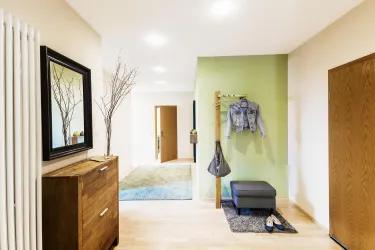 Ein moderner Flur mit einem Holzschrank und einem Spiegel. An der Wand hängen eine Jacke und eine Tasche, während ein Hocker und ein Teppich den Bereich ergänzen.