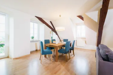 Ein heller, moderner Essbereich mit einem Holztisch und blauen Stühlen. Große Fenster sorgen für viel Tageslicht und eine einladende Atmosphäre.