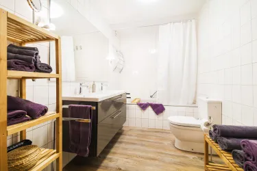 Ein modernes Badezimmer mit einem Doppelwaschbecken, einem WC und einer Badewanne in einem hellen Ambiente. Handtücher sind ordentlich gestapelt auf einem Regal aus Holz.