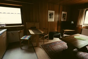 Ein gemütlicher Wohnraum mit Holzverkleidung, einem Tisch und mehreren Stühlen. Ein Fernseher steht neben einem Fenster, das Licht hereinlässt.