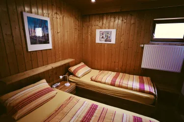 Ein gemütliches Zimmer mit zwei Betten und Holzwänden. An der Wand hängen Bilder, und es gibt ein Fenster, das für Tageslicht sorgt.