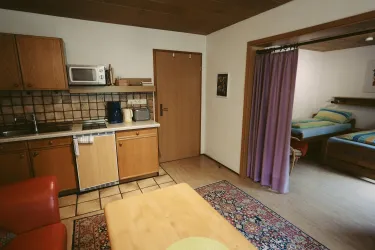 Eine gemütliche Küchenzeile mit Holzschränken und einem Tisch. Im Hintergrund sieht man ein Schlafzimmer mit zwei Betten und einem Vorhang.