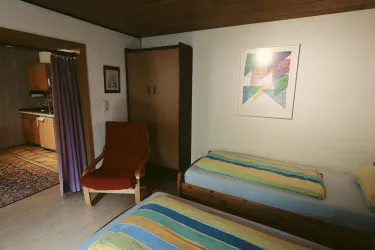 Ein einfaches Zimmer mit zwei Einzelbetten und einem roten Stuhl. Im Hintergrund sind eine Küche und ein Kleiderschrank zu sehen.