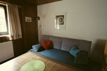 Ein gemütlicher Wohnraum mit einer Couch, dekorativen Kissen und einem Tisch. An der Wand hängt ein Bild, und das Fenster lässt sanftes Licht herein.