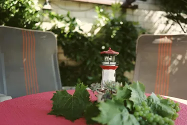 Ein Tisch mit einer pinken Decke, auf dem ein kleiner Leuchtturm steht. Umgeben von grünen Pflanzen und etwas Trauben.