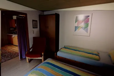 Ein einfaches Zimmer mit zwei Betten und bunten Bettdecken. Im Hintergrund ist eine Küche und ein Kunstwerk an der Wand zu sehen.