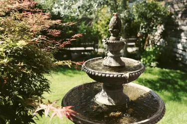 Ein schöner Brunnen im Garten mit sprudelndem Wasser und umgeben von buntem Grün. Die Szene strahlt Ruhe und Naturverbundenheit aus.