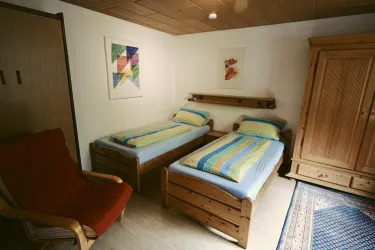 Ein gemütliches Schlafzimmer mit zwei Einzelbetten und bunten Bettdecken. An der Wand hängen mehrere Bilder, und ein roter Sessel steht in der Ecke.