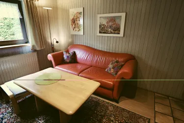 Ein gemütliches Wohnzimmer mit einem roten Sofa und einem Holzkaffee-Tisch. An den Wänden hängen zwei Bilder, und der Boden ist mit einem bunten Teppich bedeckt.