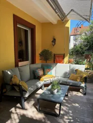 Eine einladende Terrasse mit einer gemütlichen Sitzgarnitur und bunten Kissen. Die Wände sind in warmen Gelbtönen gestrichen, umgeben von Pflanzen.