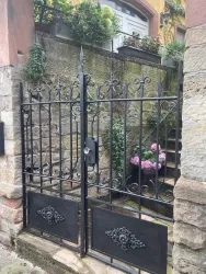 Ein schmiedeeisernes Tor mit geschwungenen Verzierungen führt zu einem kleinen Garten. Im Hintergrund sind blühende Pflanzen und Treppen sichtbar.