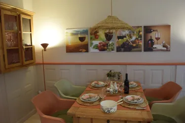 Ein einladender Essbereich mit einem Holztisch und bunten Stühlen. An der Wand hängen Bilder mit Wein und frischem Obst.