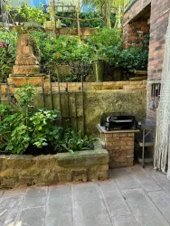 Ein kleiner Garten mit üppigem Grün und einer schönen Steinmauer. Im Vordergrund steht ein Grill auf einem Tisch.
