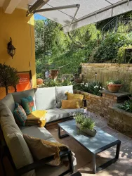 Eine gemütliche Terrasse mit bequemen Sitzgelegenheiten und einem kleinen Tisch. Im Hintergrund sind grüne Pflanzen und eine Wand mit Blumen zu sehen.