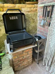Ein tragbarer Grill steht auf einem Steinsockel in einem Außenbereich. Neben dem Grill sind einige Grillutensilien aufgehängt.