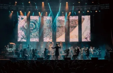 Ein Orchester spielt auf einer Bühne, während ein Dirigent das Ensemble leitet. Im Hintergrund sind große Leinwände mit bunten Mustern und Lichtern zu sehen.