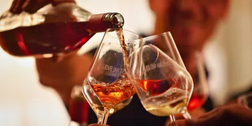 Ein paar Gläser werden gefüllt und stoßen an. Im Hintergrund sieht man eine fröhliche Atmosphäre.