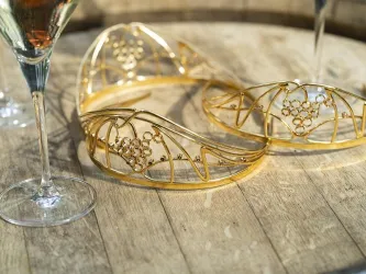 Zwei elegante, goldene Haarschmuckstücke liegen auf einem Holztablett. Im Hintergrund stehen Gläser mit Schaumwein.