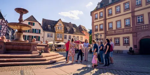 Eine Gruppe von Menschen steht an einem Brunnen auf einem malerischen Platz. Im Hintergrund sind schöne, traditionelle Gebäude und ein blauer Himmel zu sehen.