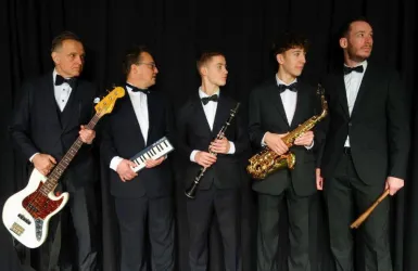 Eine Gruppe von fünf Musikern steht nebeneinander. Sie tragen elegante Anzüge und halten verschiedene Musikinstrumente.