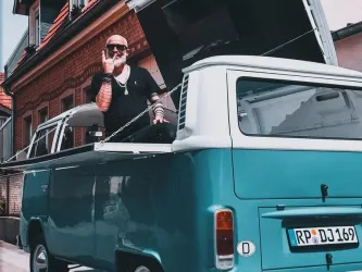 Ein Mann sitzt in einem grünen VW-Bus und zeigt das Rock-Zeichen. Der Hintergrund zeigt ein typisches Stadtbild mit Häusern.