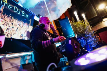 Ein DJ steht an einem Mischpult und spielt Musik vor einer begeisterten Menschenmenge. Die Atmosphäre ist lebhaft mit bunten Lichtern im Hintergrund.