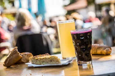 Ein Tisch mit zwei Getränken, einem Bier und einer dunklen Limonade, sowie verschiedenen Backwaren. Im Hintergrund sind unscharfe Personen und eine sonnige Atmosphäre zu sehen.