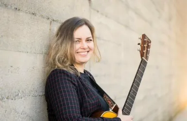 Eine Frau mit langen, welligen Haaren lächelt und steht an einer Wand. Sie hält eine Gitarre und trägt ein schwarzes, kariertes Outfit.