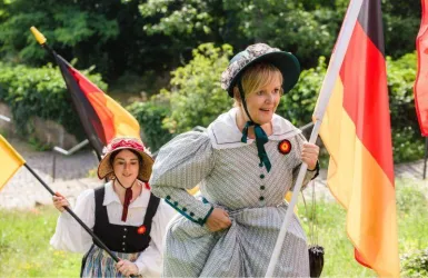 Zwei Frauen in historischen Kostümen tragen deutsche Fahnen. Sie gehen eine Grünfläche entlang und präsentieren damit einen festlichen Anlass.