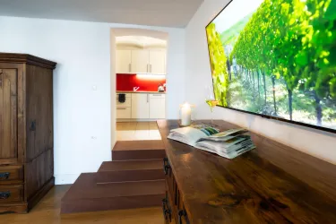 Ein heller Wohnraum mit Holzschränken und einem modernen Fernseher. Im Hintergrund ist eine Küche mit roten Akzenten sichtbar.