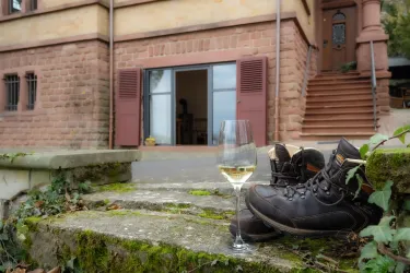 Ein Paar Wanderschuhe steht neben einem Glas Weißwein auf einer steinernen Treppe. Im Hintergrund ist ein Gebäude mit offenen Fenstern zu sehen.