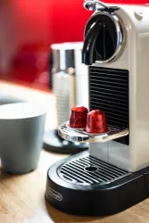 Eine Nespresso-Maschine mit zwei roten Kaffeekapseln steht auf einer Holzoberfläche. Im Hintergrund sind eine Tasse und eine Milchkanne zu sehen.