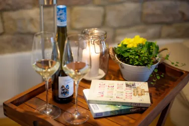 Auf einem Tablett stehen zwei Weingläser, eine Flasche Wein und ein Glas mit einer Kerze. Daneben liegen zwei Bücher und ein Topf mit grünen Pflanzen.