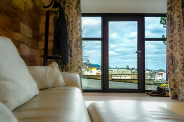 Ein gemütlicher Raum mit einem weißen Sofa und Blick auf eine malerische Landschaft. Draußen sind Bäume und Gebäude sichtbar, unter einem bewölkten Himmel.