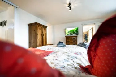 Ein gemütliches Schlafzimmer mit einem großen Bett und einer roten Bettdecke. Im Hintergrund sind ein Holzschrank und ein Fernseher sichtbar.