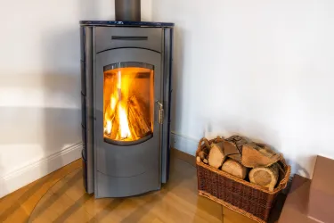 Ein moderner Kaminofen mit flammendem Feuer steht in einem gemütlichen Raum. Neben dem Ofen steht ein Korb mit Holzscheiten zur Verfügung.