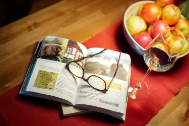 Ein offenes Buch liegt auf einem Tisch, daneben liegen eine Brille und ein Glas Wein. Im Hintergrund steht eine Schüssel mit bunten Äpfeln.