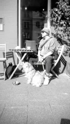 Eine Person sitzt an einem Tisch im Freien, neben einem kleinen Hund. Es ist sonnig und die Atmosphäre ist entspannt.