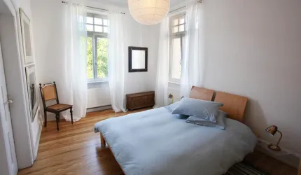 Ein helles Schlafzimmer mit einem großen Bett und blauen Kissen. Große Fenster bringen viel Tageslicht in den Raum.