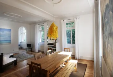 Ein heller und geräumiger Essbereich mit einem großen Holztisch und Stühlen. An der Wand hängt ein gemaltes Bild, und es gibt große Fenster mit Blick ins Grüne.