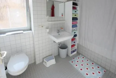 Ein helles Badezimmer mit weißen Fliesen und einem Waschbecken. Neben dem Waschbecken steht ein Regal mit bunten Aufbewahrungsboxen.