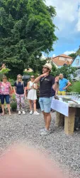 Eine Person spricht mit einer Gruppe von Menschen im Freien. Im Hintergrund sind Tische mit Getränken und eine grüne Umgebung zu sehen.