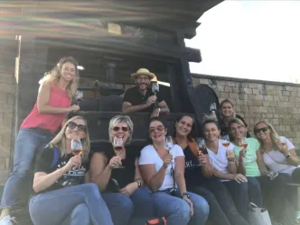 Eine Gruppe von Frauen posiert fröhlich mit Weingläsern. Im Hintergrund ist eine alte Weinpresse sichtbar.
