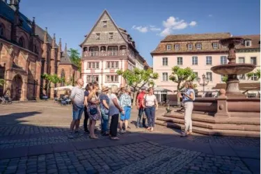 Eine Gruppe von Touristen hört einem Führer auf einem Platz in einer Stadt zu. Im Hintergrund sind traditionelle Gebäude und ein Brunnen sichtbar.