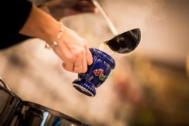 Eine Hand gießt heißen Inhalt mit einem Löffel in einen bunten Becher. Dampfwolken steigen auf und verleihen der Szene eine warme Atmosphäre.