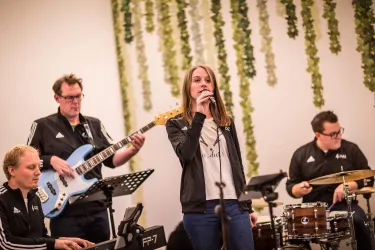 Eine Band spielt live auf einer Bühne. Eine Sängerin steht im Vordergrund und wird von Musikern an Gitarre und Schlagzeug begleitet.